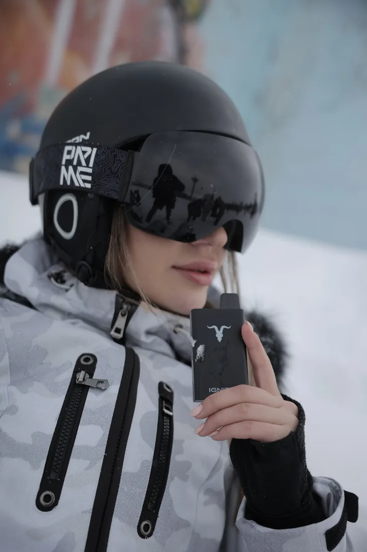 Skier with a black helmet and goggles holding a black IGNITE device, snow in the background