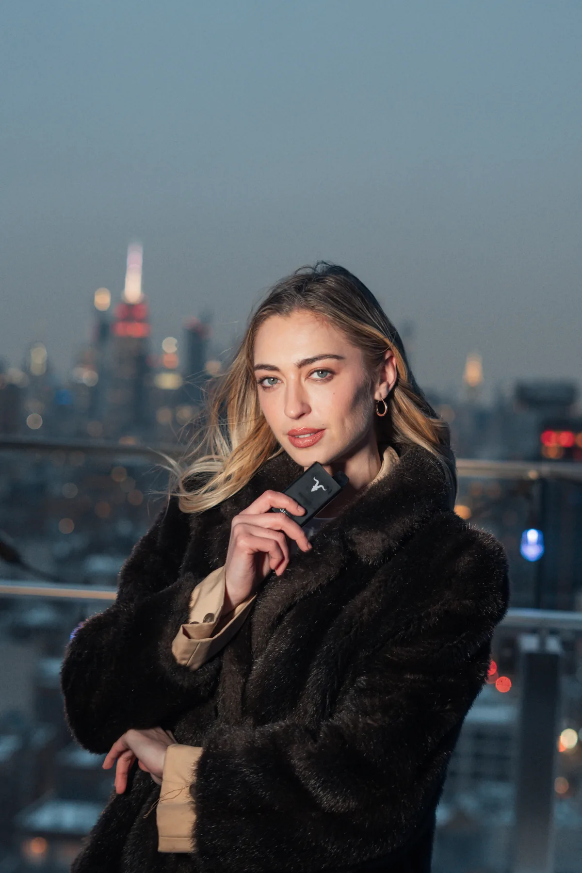 Woman in a dark coat holding an IGNITE device on a rooftop, city skyline at dusk behind her