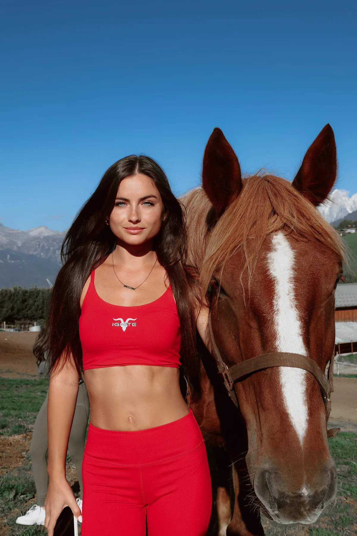 Woman in a red IGNITE sports outfit next to a horse in front of mountains